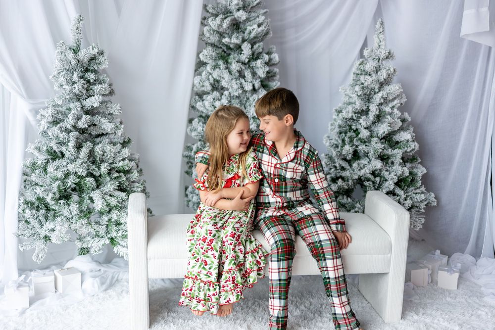 Two children in Christmas pajamas sit together on a white bench surrounded by snow-flocked holiday trees.