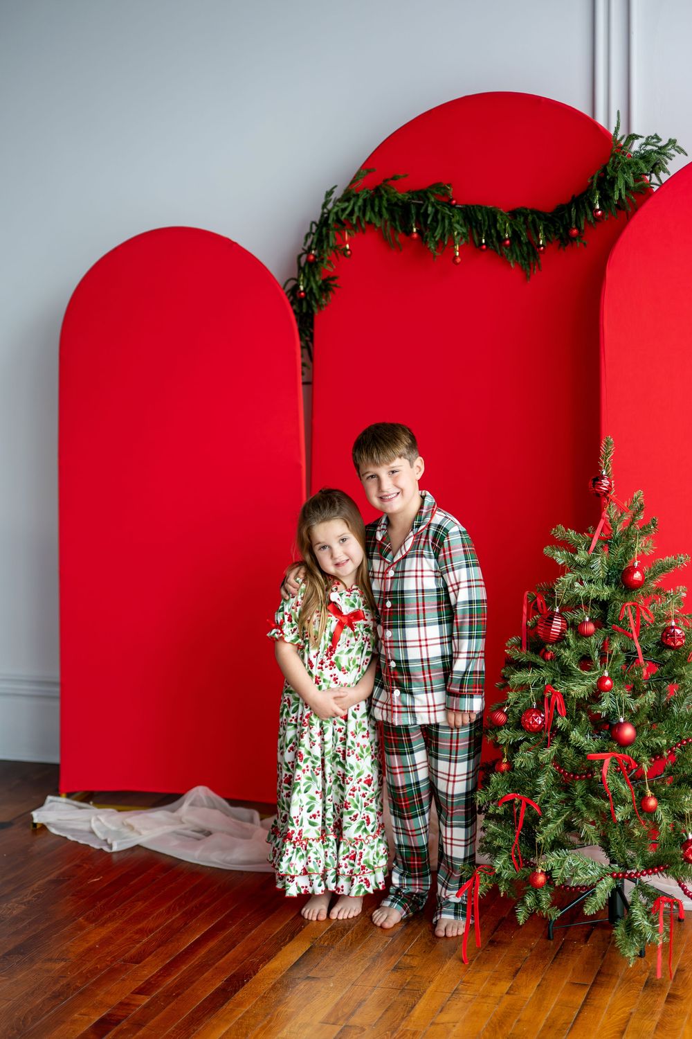 A boy and girl in matching Christmas pajamas pose together beside a decorated tree and red backdrop.