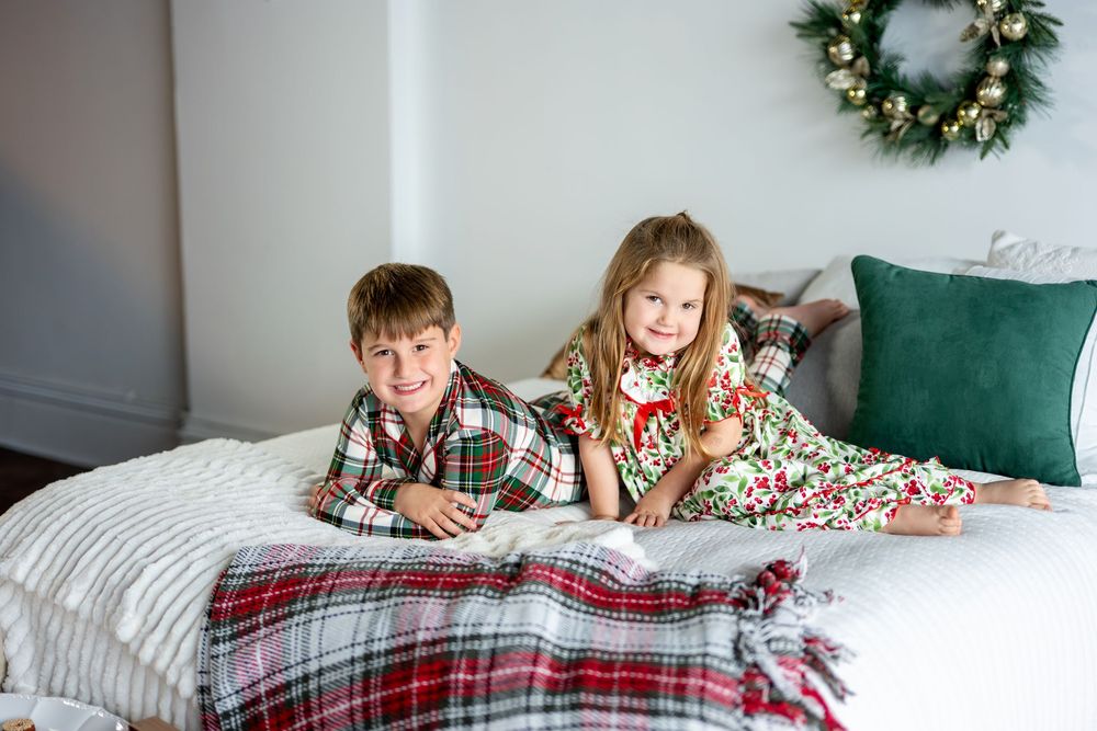 Two children in festive Christmas pajamas lie on a bed with a plaid blanket and holiday wreath decor.