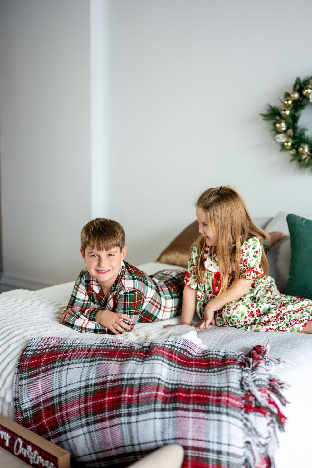 A boy in plaid Christmas pajamas lies on a bed while his sister sits beside him near a holiday wreath.
