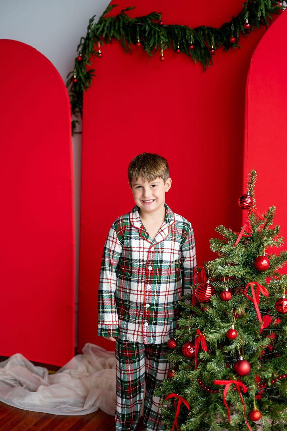 A smiling boy in plaid Christmas pajamas stands next to a decorated tree against a bold red backdrop.
