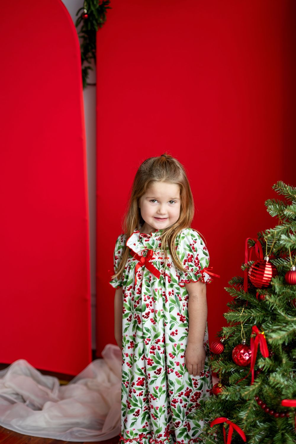 A young girl in a white and green holly-print Christmas dress smiles beside a decorated red holiday tree.
