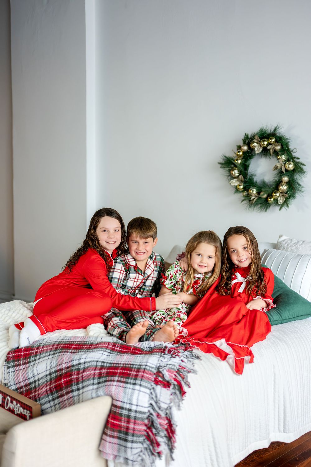 Four children in Christmas pajamas sit together smiling on a bed decorated with a holiday wreath.
