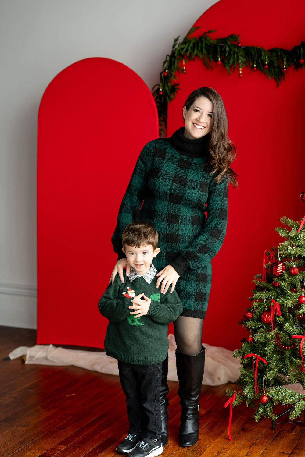 A smiling mother in green plaid stands with her young son beside a decorated Christmas tree and red arch photo backdrop.