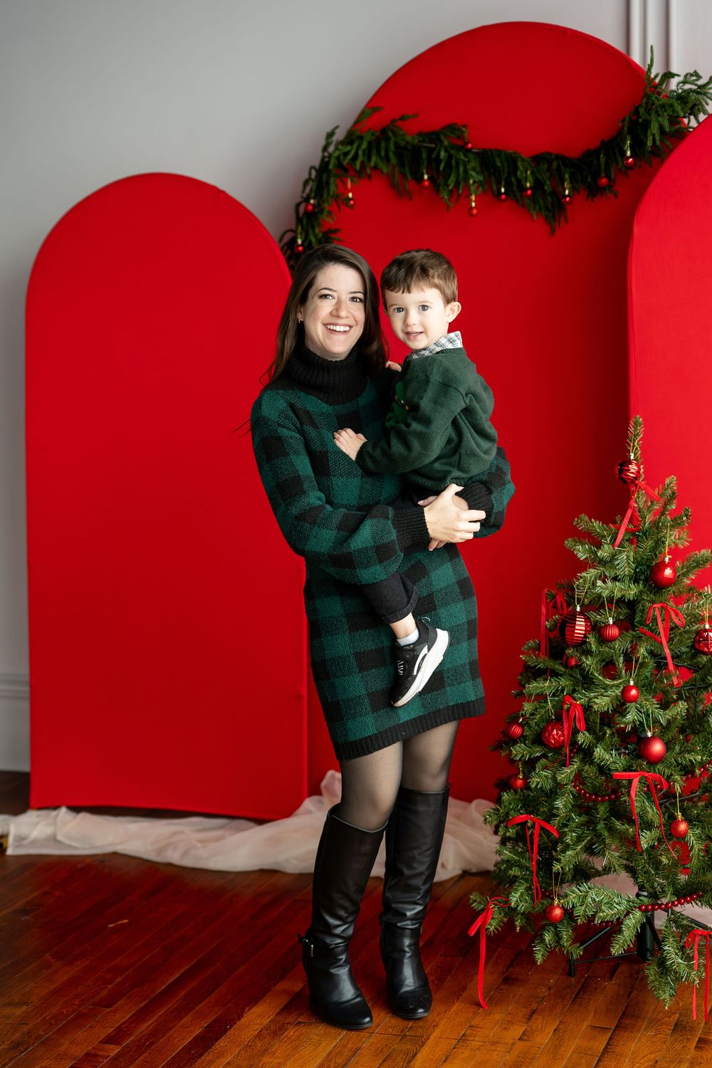 A joyful mother in a green plaid dress holds her young son in her arms beside a decorated Christmas tree and red backdrop.