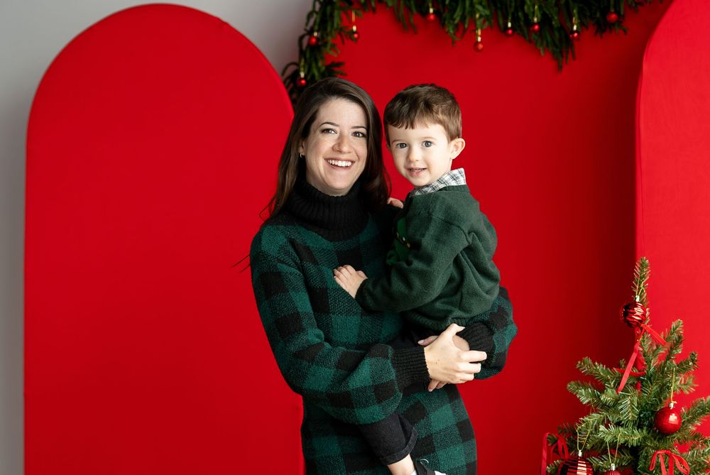 A smiling mother in a green plaid turtleneck holds her young son in front of red arch backdrops and a Christmas tree.