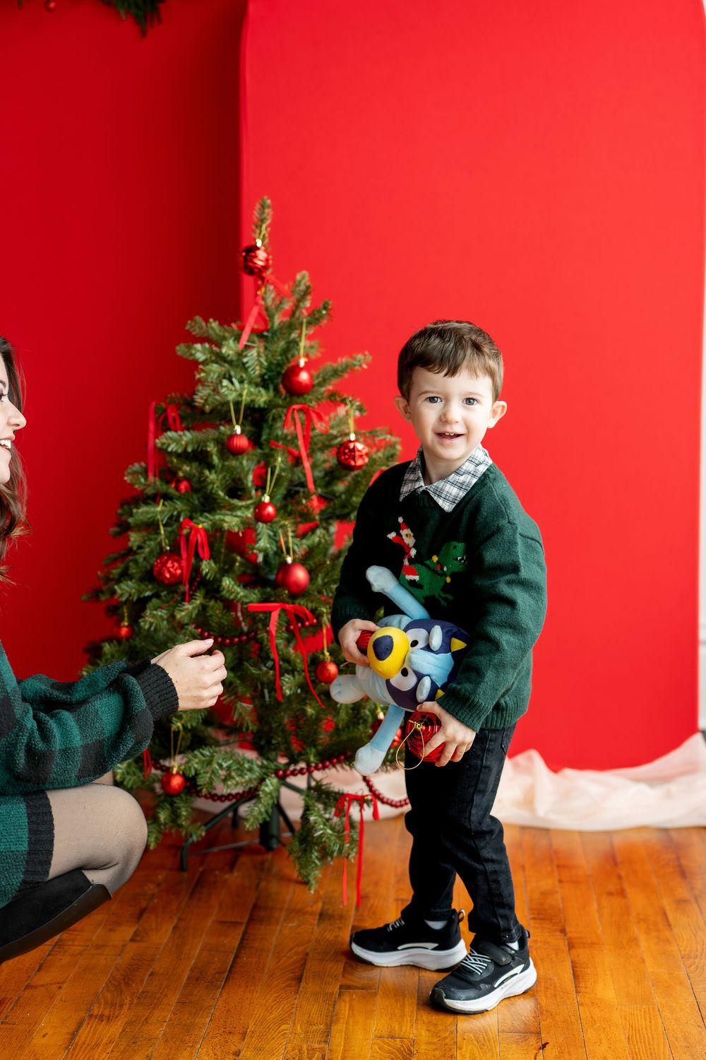 Young boy in green Christmas sweater holding Bluey toy beside decorated Christmas tree.