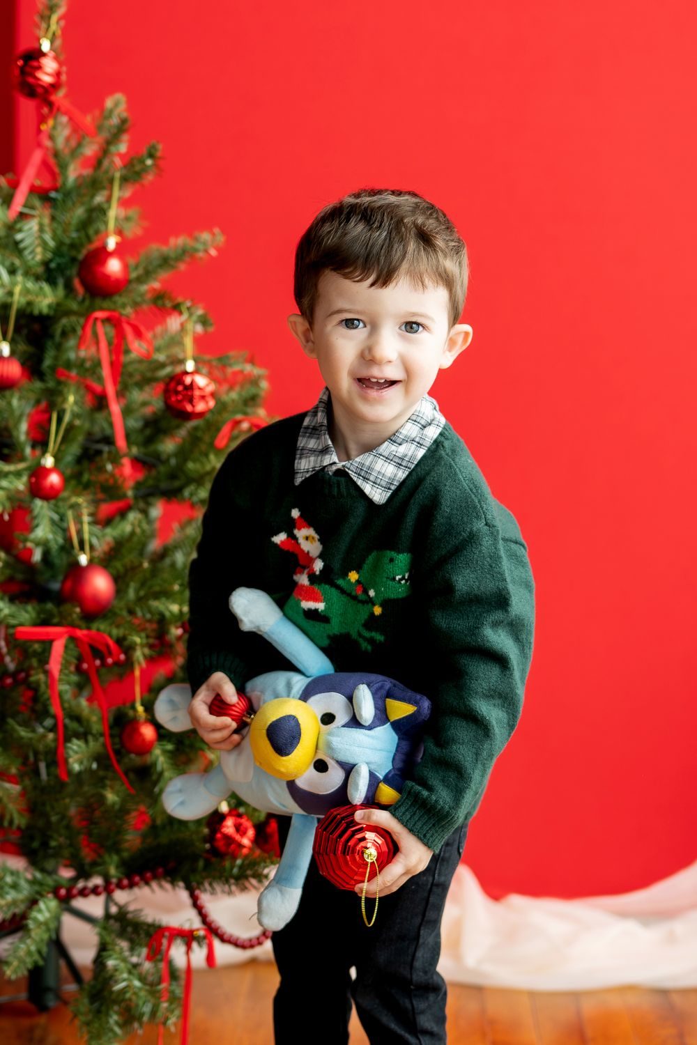 Smiling toddler boy in green reindeer sweater holding Bluey stuffed toy by Christmas tree.