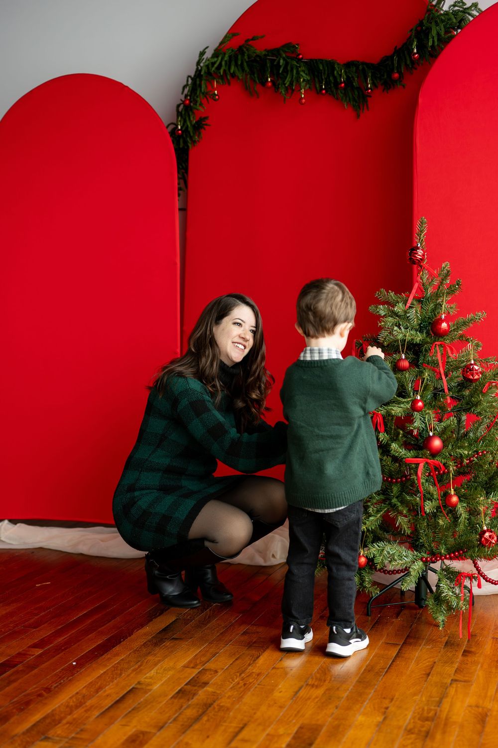 A smiling mother in a green plaid dress crouches beside her young son as they decorate a Christmas tree with red ornaments.