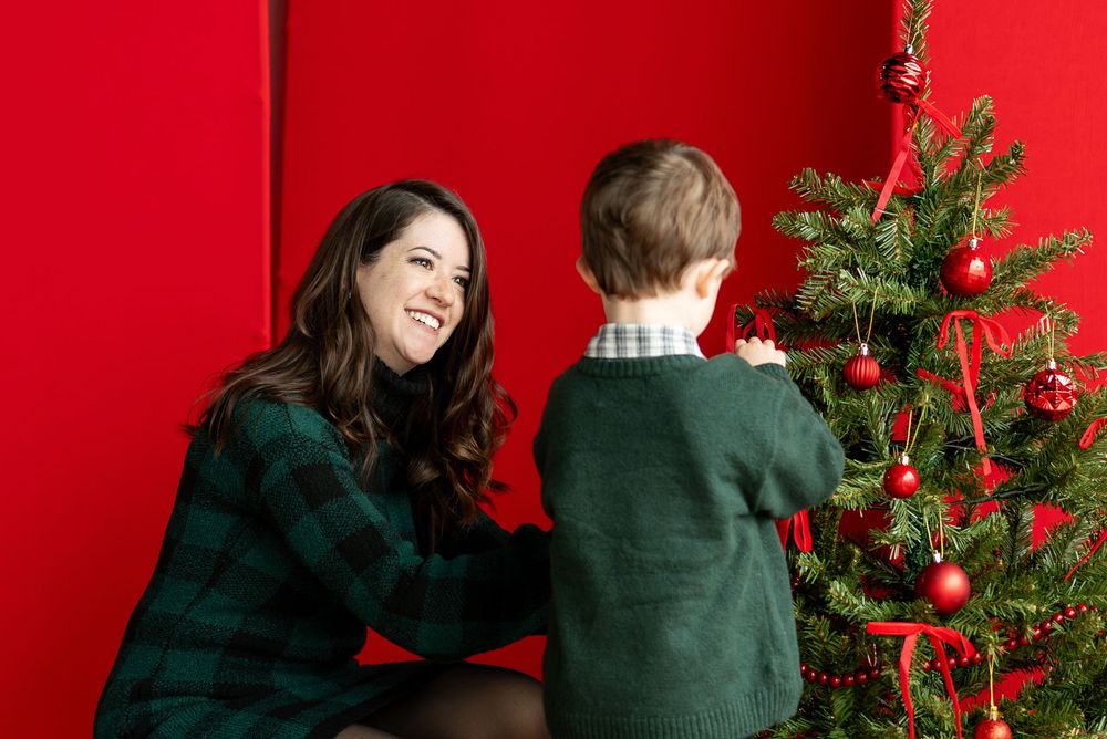 A laughing mother in green plaid sits with her young son while he touches ornaments on a red-decorated Christmas tree.