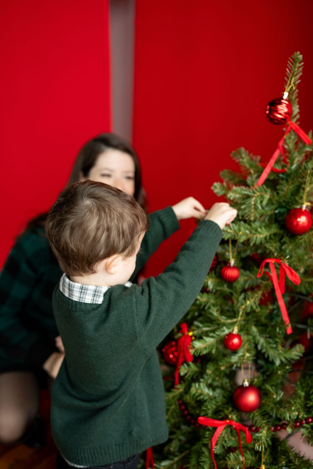A young boy in a green sweater reaches up to place a red ornament on a Christmas tree while his mother assists from behind.