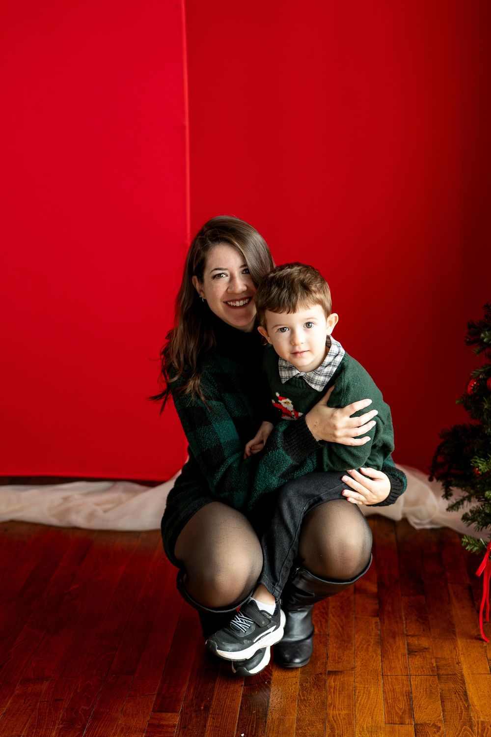 A smiling mother in a green plaid dress crouches on a wood floor hugging her young son in front of a red backdrop.