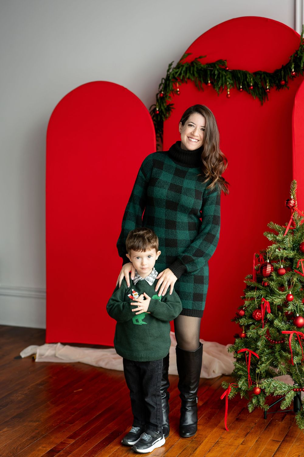 A mother in a green plaid dress stands with her young son holding a toy in front of red arch backdrops and a Christmas tree.