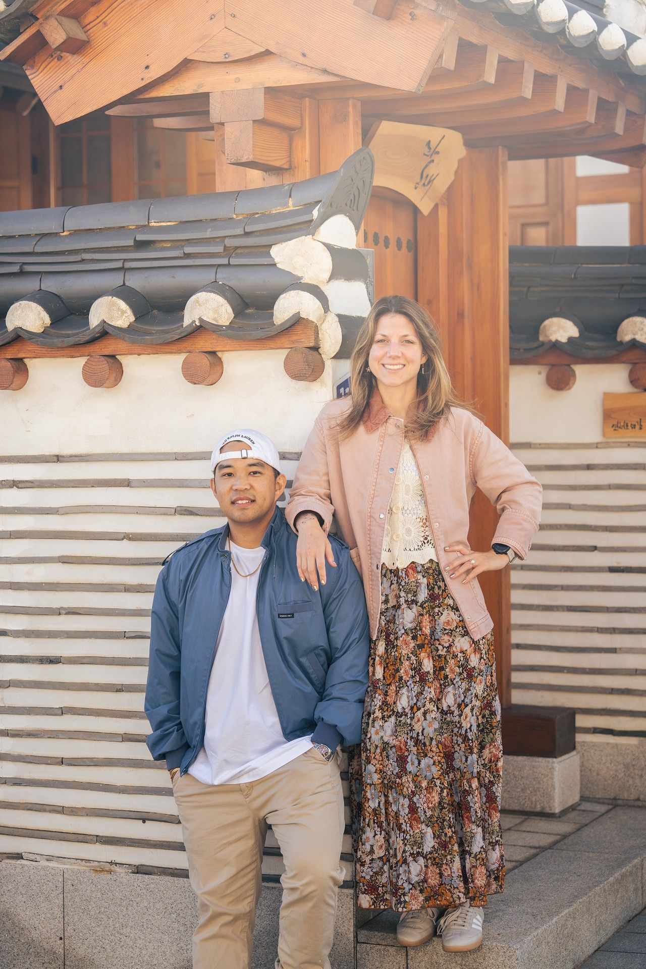 A couple poses together in traditional Korean hanbok clothing outside a temple with wooden architecture and stone walls.