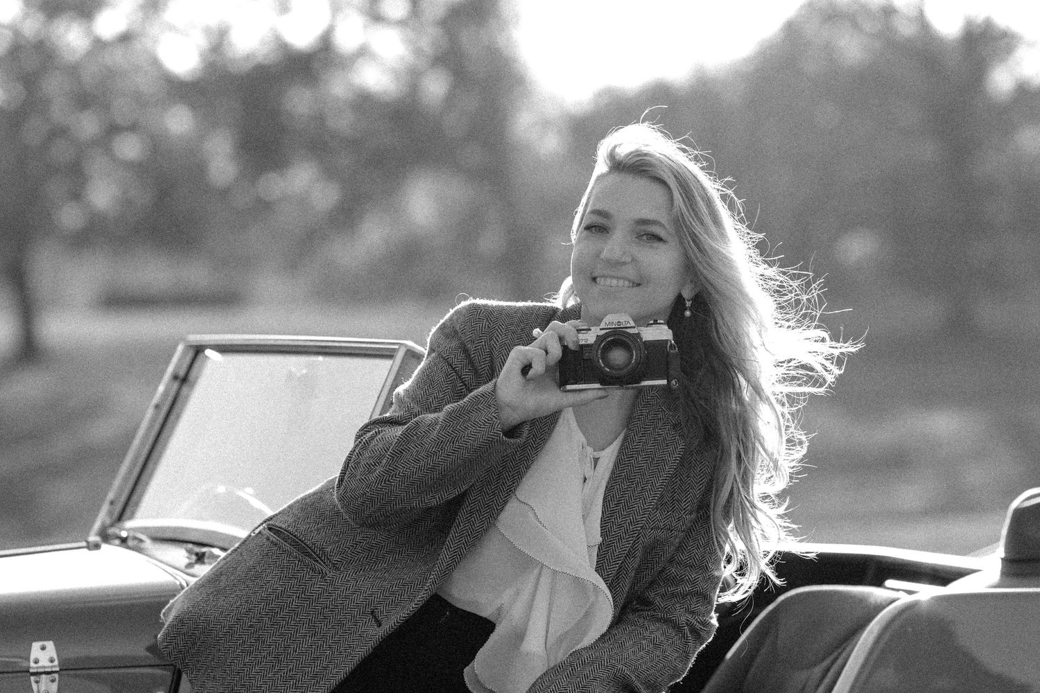 Smiling blonde woman holding vintage camera in convertible car in black and white.