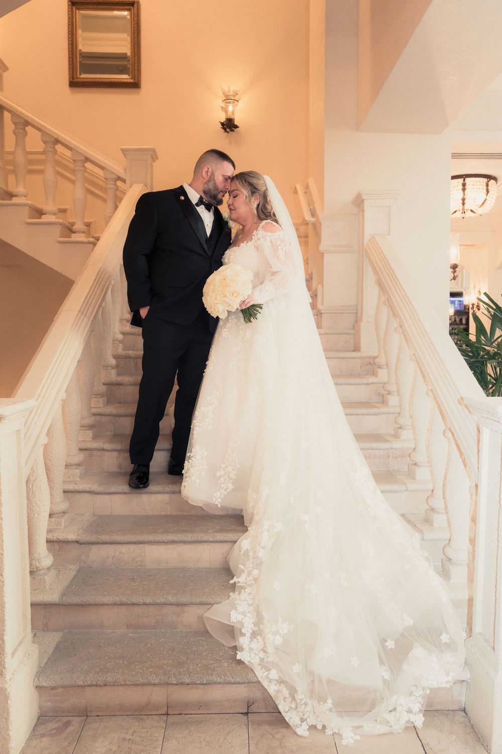 Bride and groom share a romantic moment on elegant white marble staircase, bride wearing lace gown with long veil.
