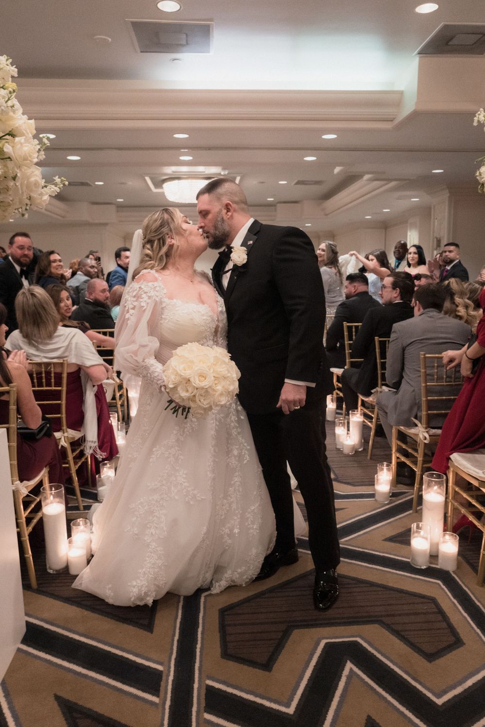Bride and groom share a kiss at the altar during their elegant indoor wedding ceremony with candles lining the aisle.