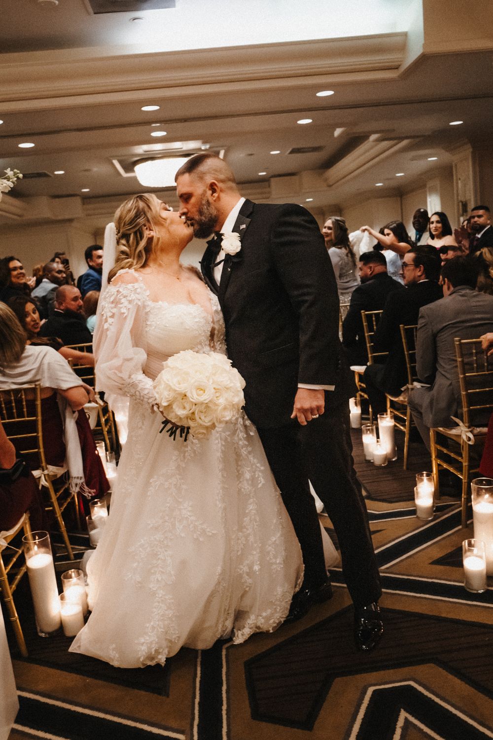 A romantic wedding moment captured in a candlelit ballroom with a bride in lace gown and groom in black tuxedo.