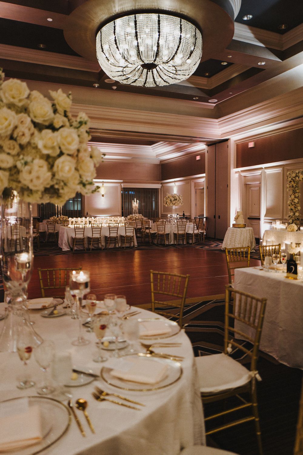 Elegant wedding reception ballroom with crystal chandeliers, white floral centerpieces, and round tables set for dinner service.