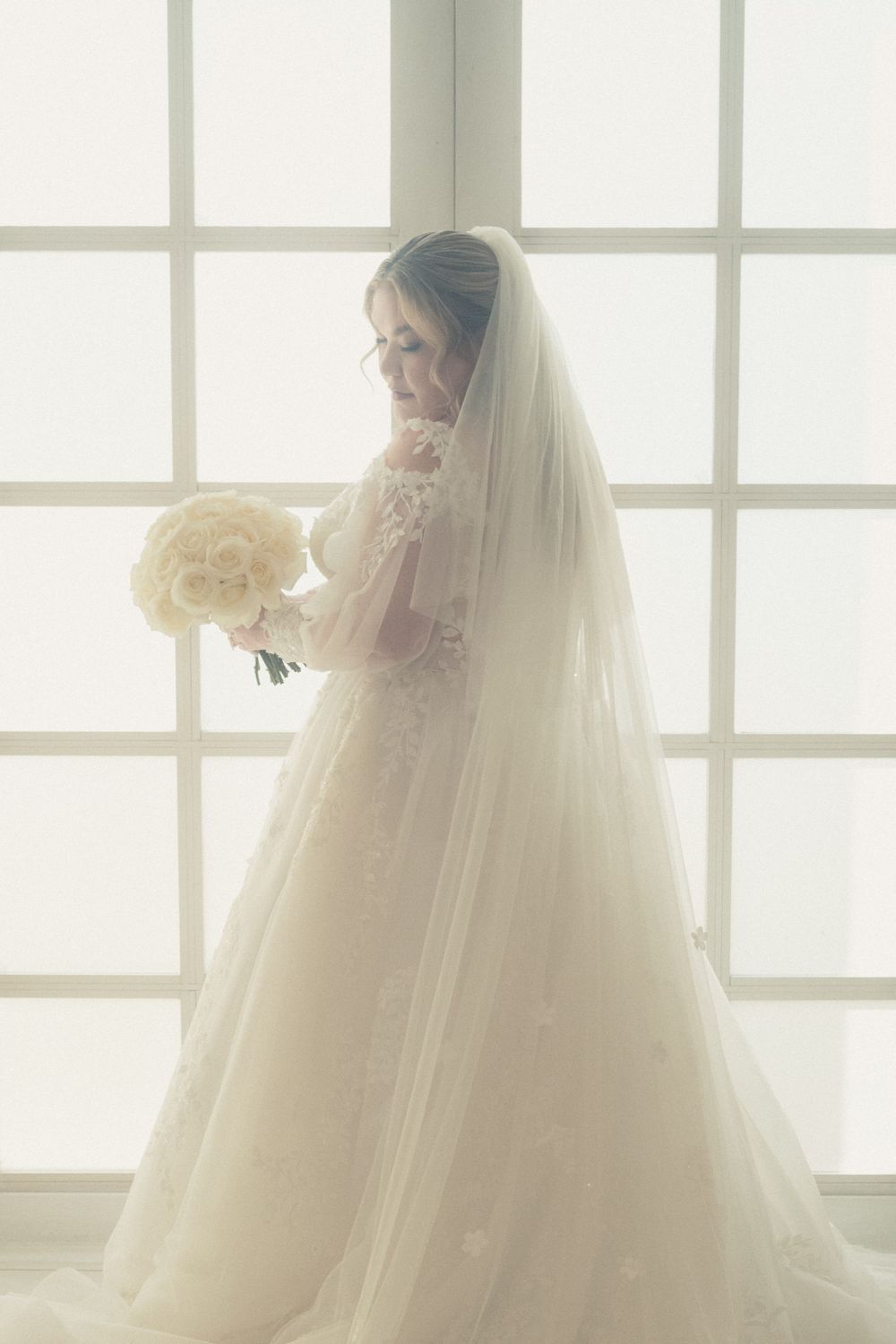 Bride in white lace gown and veil holds bouquet of white roses, standing by large bright windows.