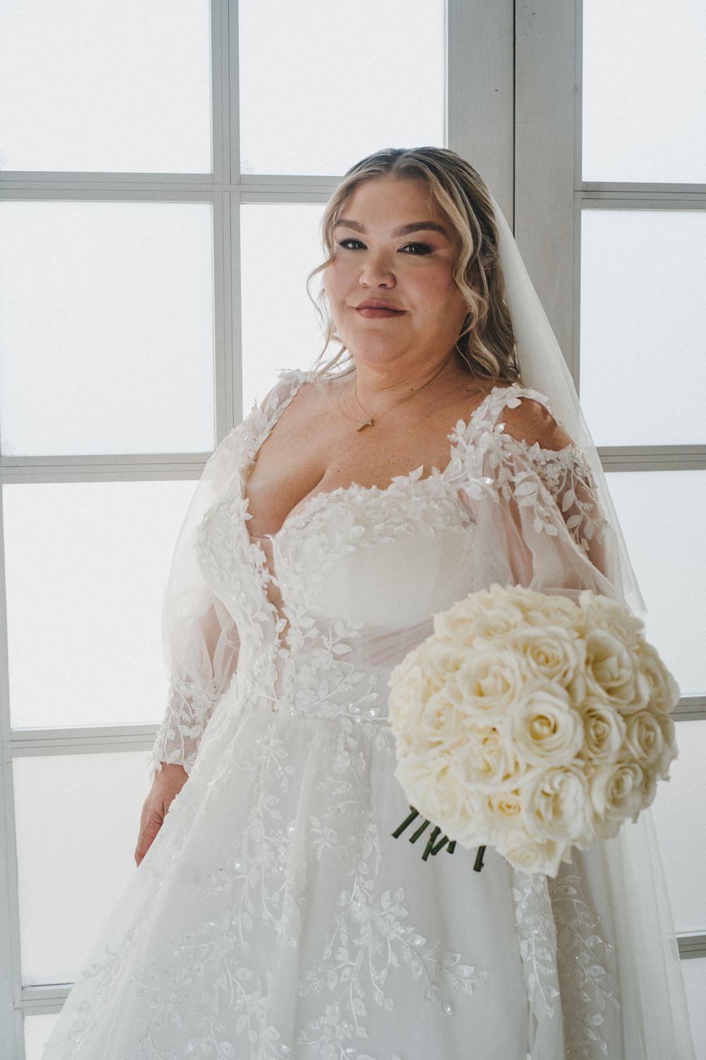A bride in a luxurious white lace wedding gown holds an elegant bouquet of cream roses against a bright window backdrop.