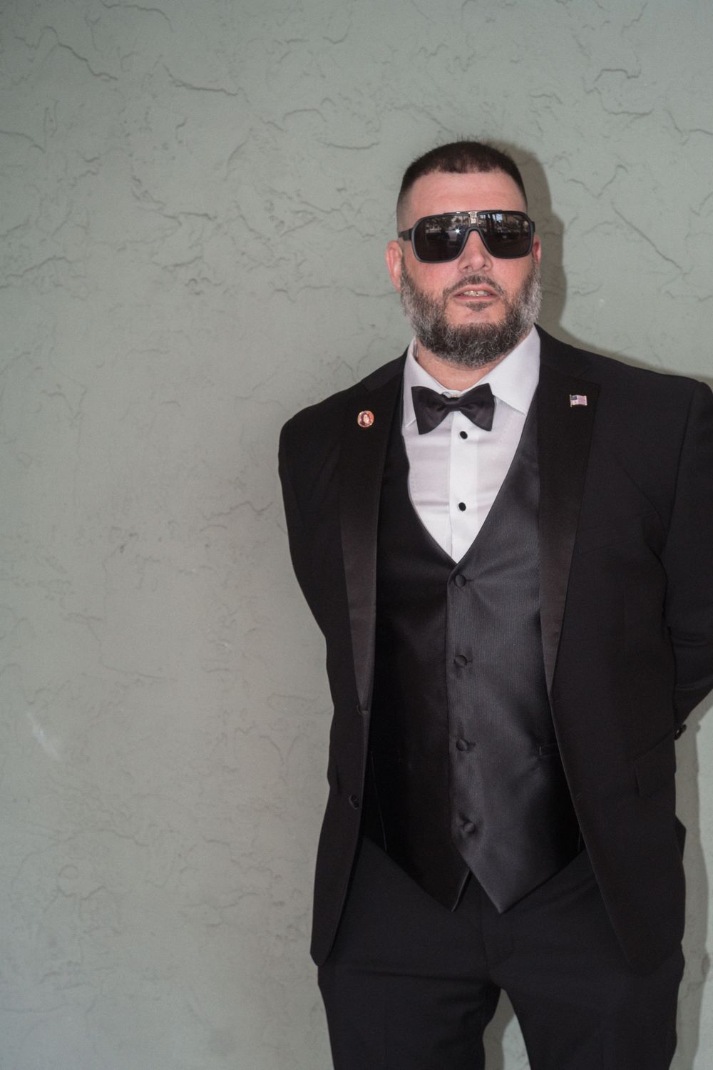 Confident groom in black tuxedo with bow tie and sunglasses posing against textured wall.