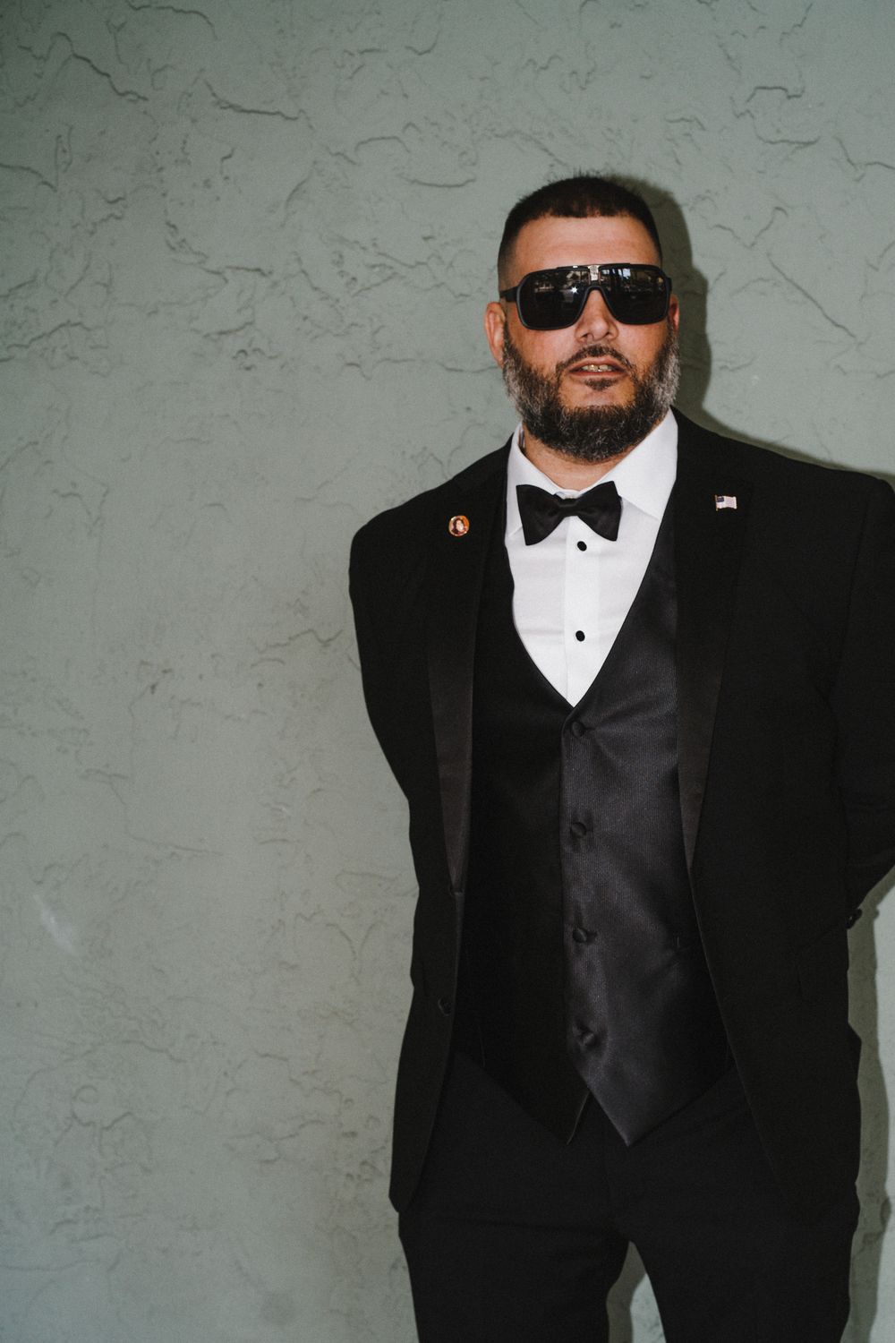 A groom poses in a classic black tuxedo with bow tie and sunglasses against a textured grey wall.