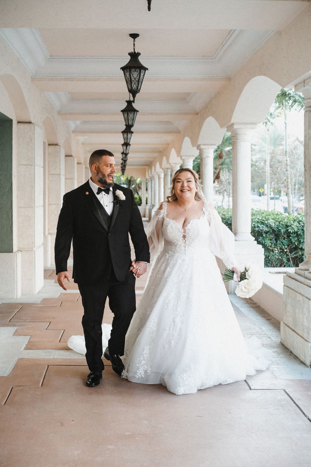 A bride and groom walk together along an elegant covered colonnade with hanging lanterns and white pillars.