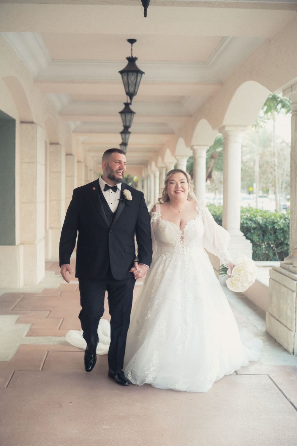 Bride and groom walk hand-in-hand under elegant arched colonnade, she in lace gown with bouquet, he in black tuxedo.