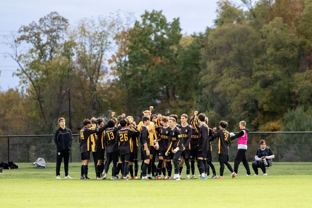 Football team in black and gold uniforms practicing on a green field during autumn season.