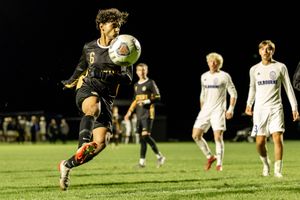 Soccer player performs a diving save during a nighttime match on a grass field while others watch in background.