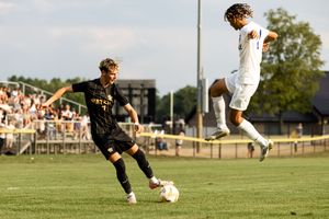 A soccer player in a white jersey jumps high in the air during a match on a sunny grass field.