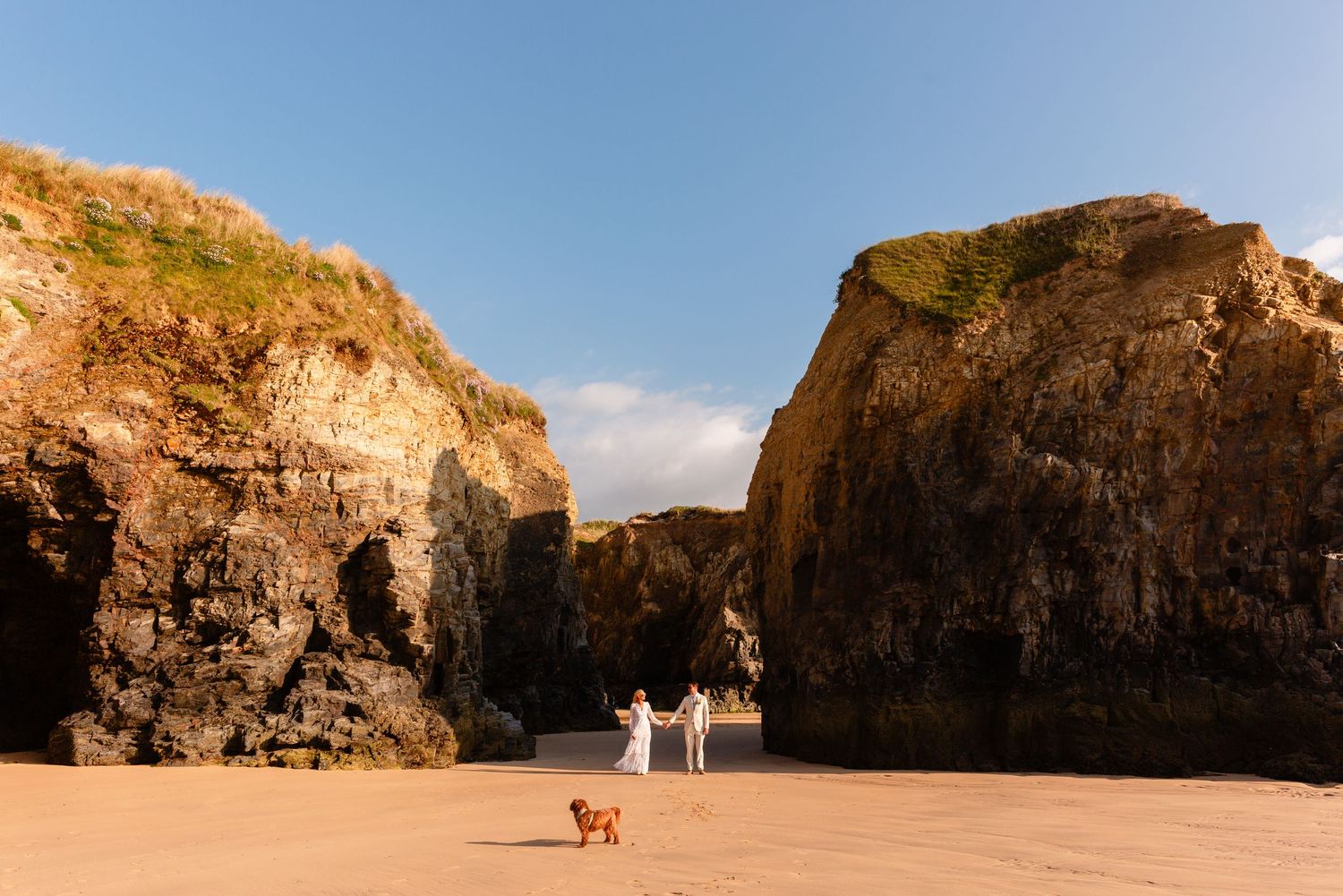 Decorative image. Wide landscape photo of the bride and groom standing between tall cliffs and outcrops along Godrevy Beach, Gwithian.