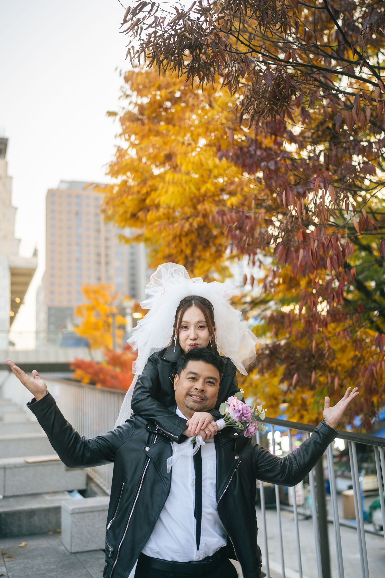 A student in a black leather jacket and white bow poses playfully against an autumn backdrop with golden trees.