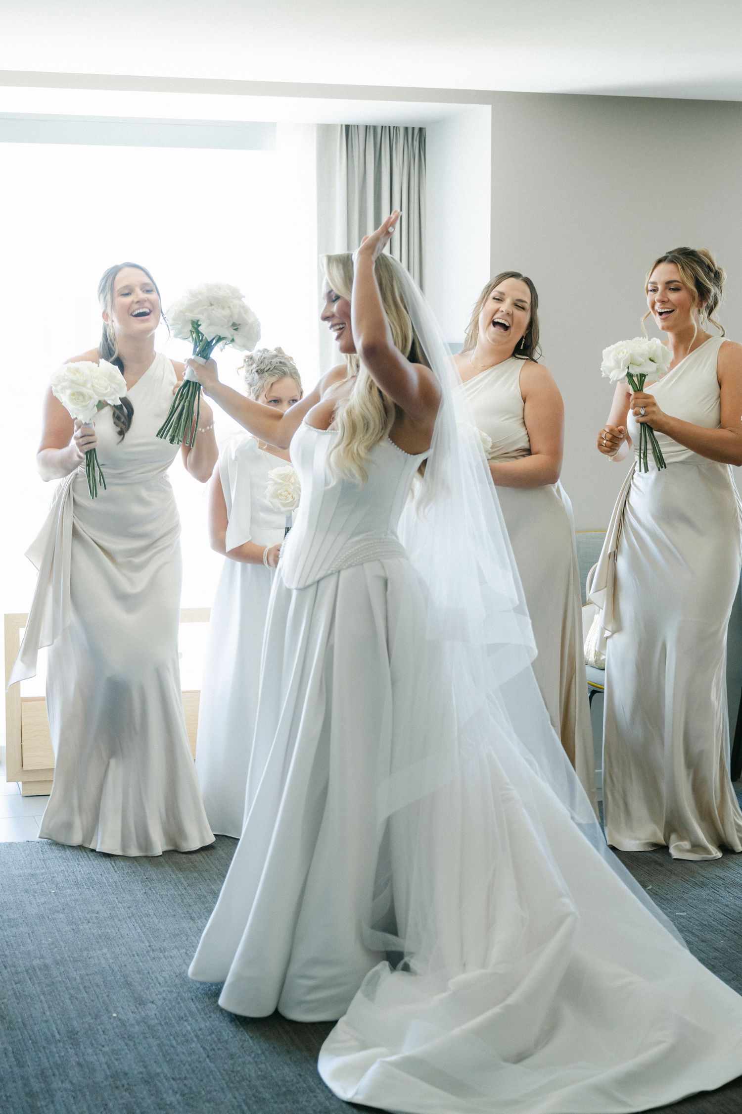 A group of bridesmaids in white dresses laugh and dance together during wedding preparations.