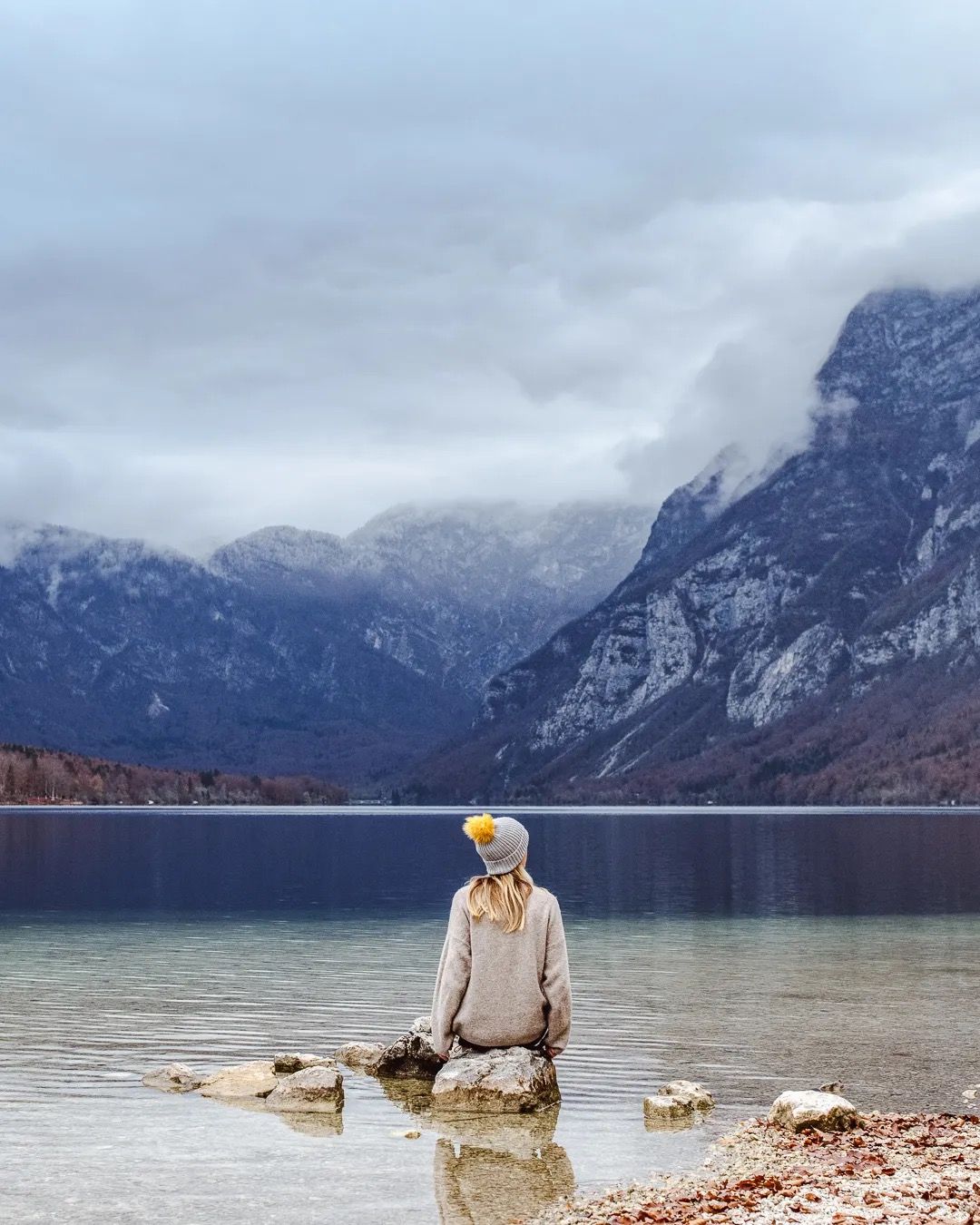 Woman standing on lakeside rocks looking at snowy mountains on a misty day.