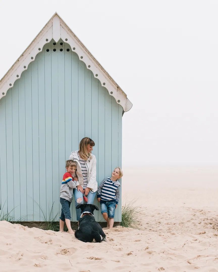 Mother and children sitting beside a pale blue beach hut on a misty beach.
