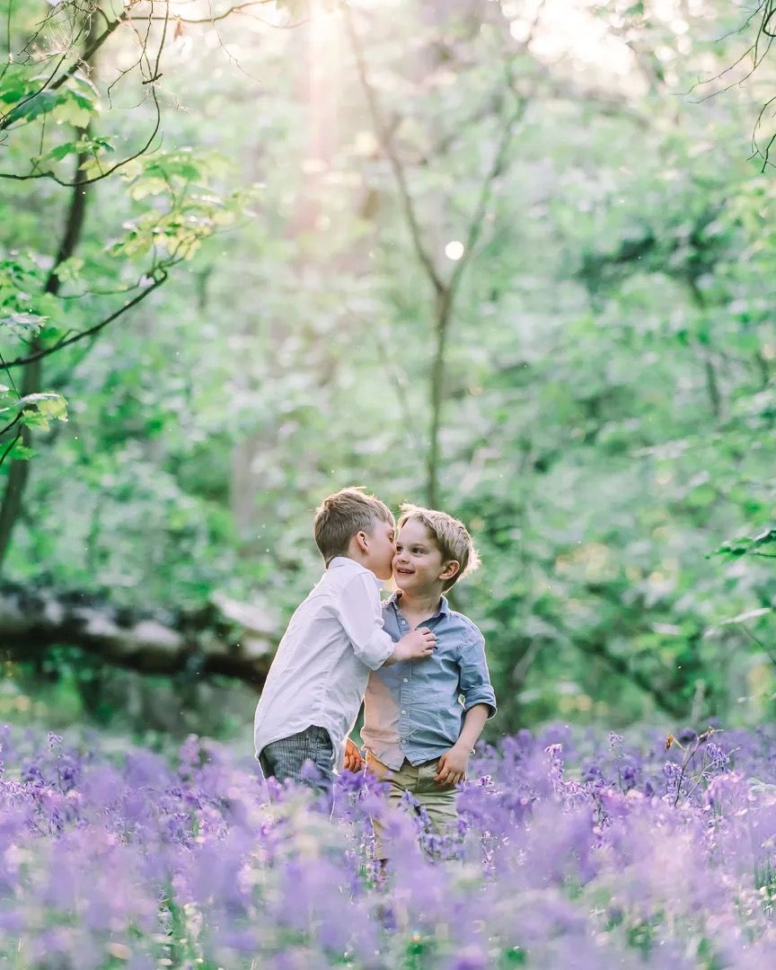 Two children whispering in a sunlit bluebell woodland with soft spring light.