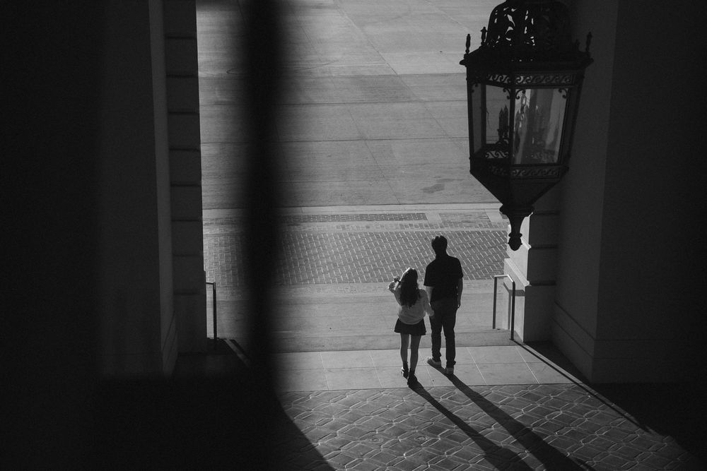 Black and white photo of couple walking under archway with long shadows at Pasadena City Hall.