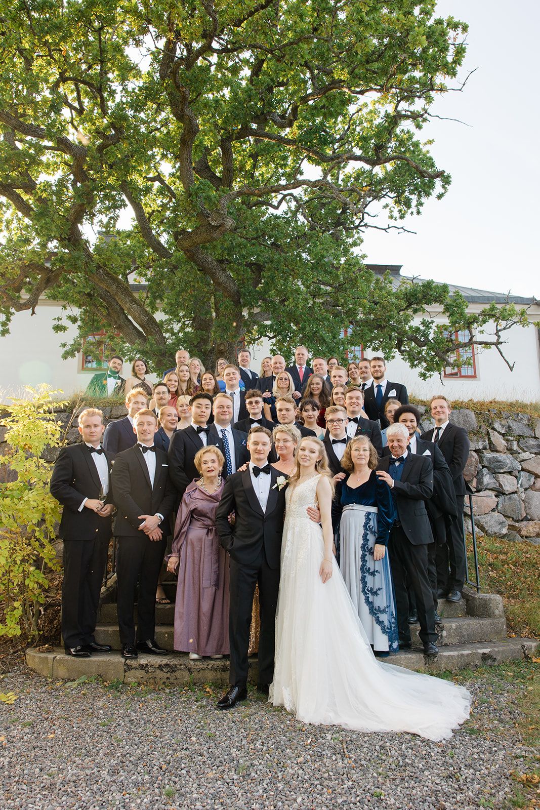 Large wedding party gathered outdoors for a formal group photo under a tree on a gravel path.