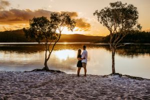 Couple standing between trees at sunset on sandy beach near tranquil lake.