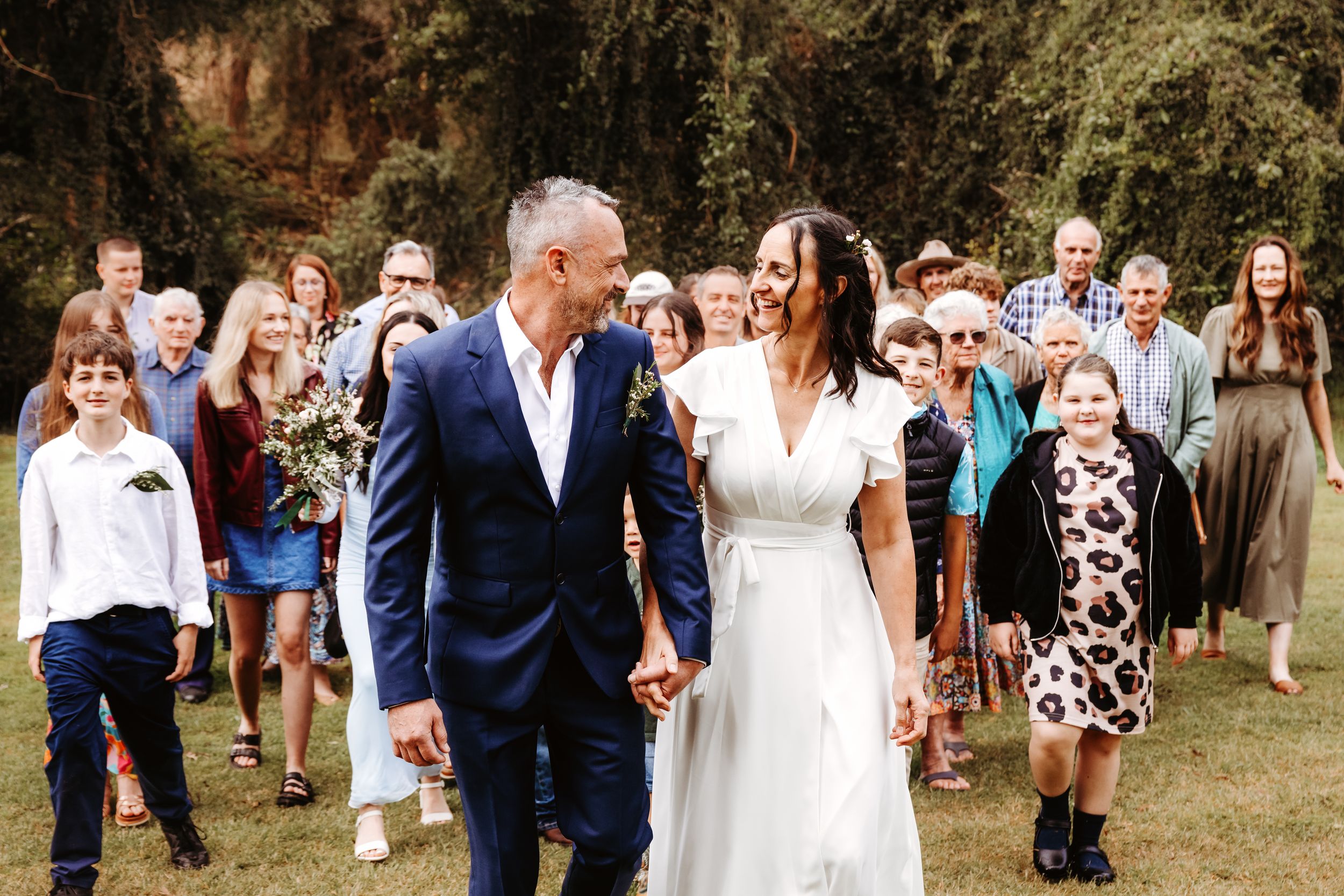 Outdoor wedding celebration with wedding party and guests walking down a grassy pathway wearing formal attire.