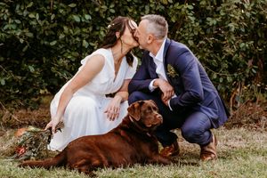 A couple shares a playful moment in a garden setting with their chocolate lab laying nearby.