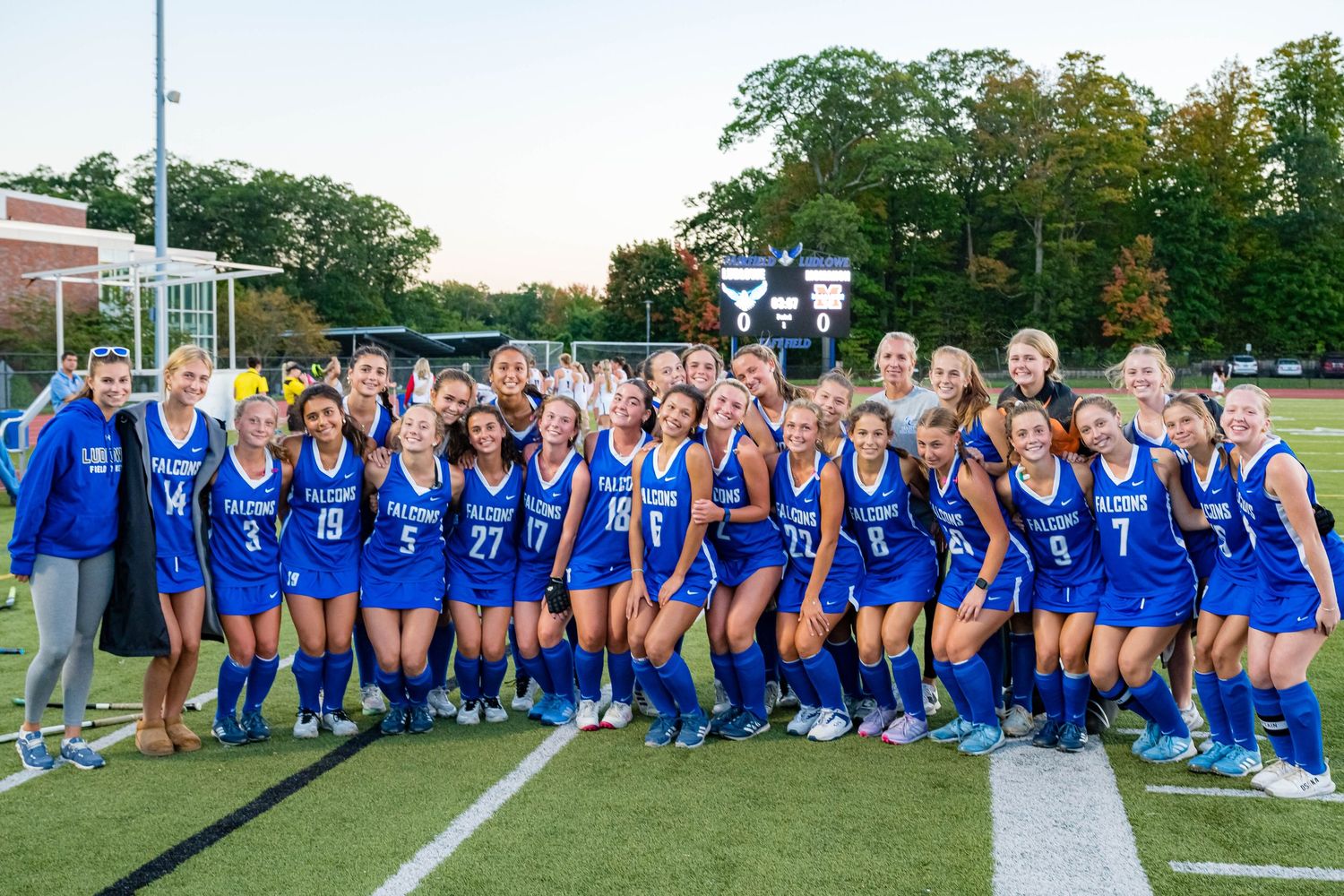 Large group of athletes in blue uniforms pose together on a sports field during a sunny day.