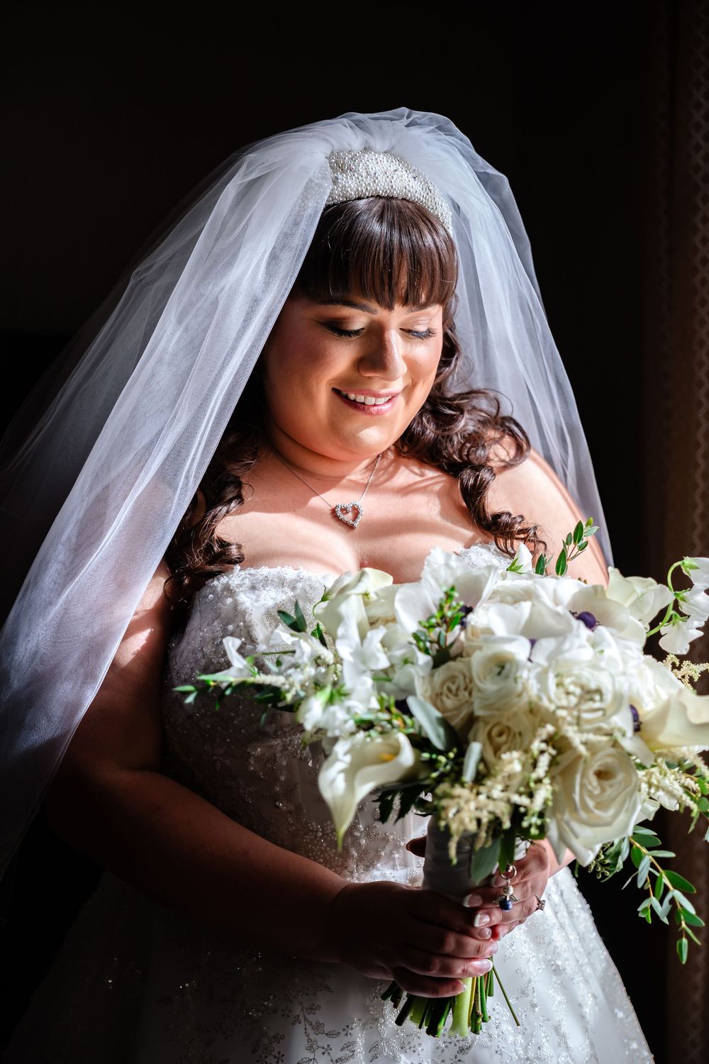A bride in a white lace gown and veil holds a bouquet of white roses and greenery.