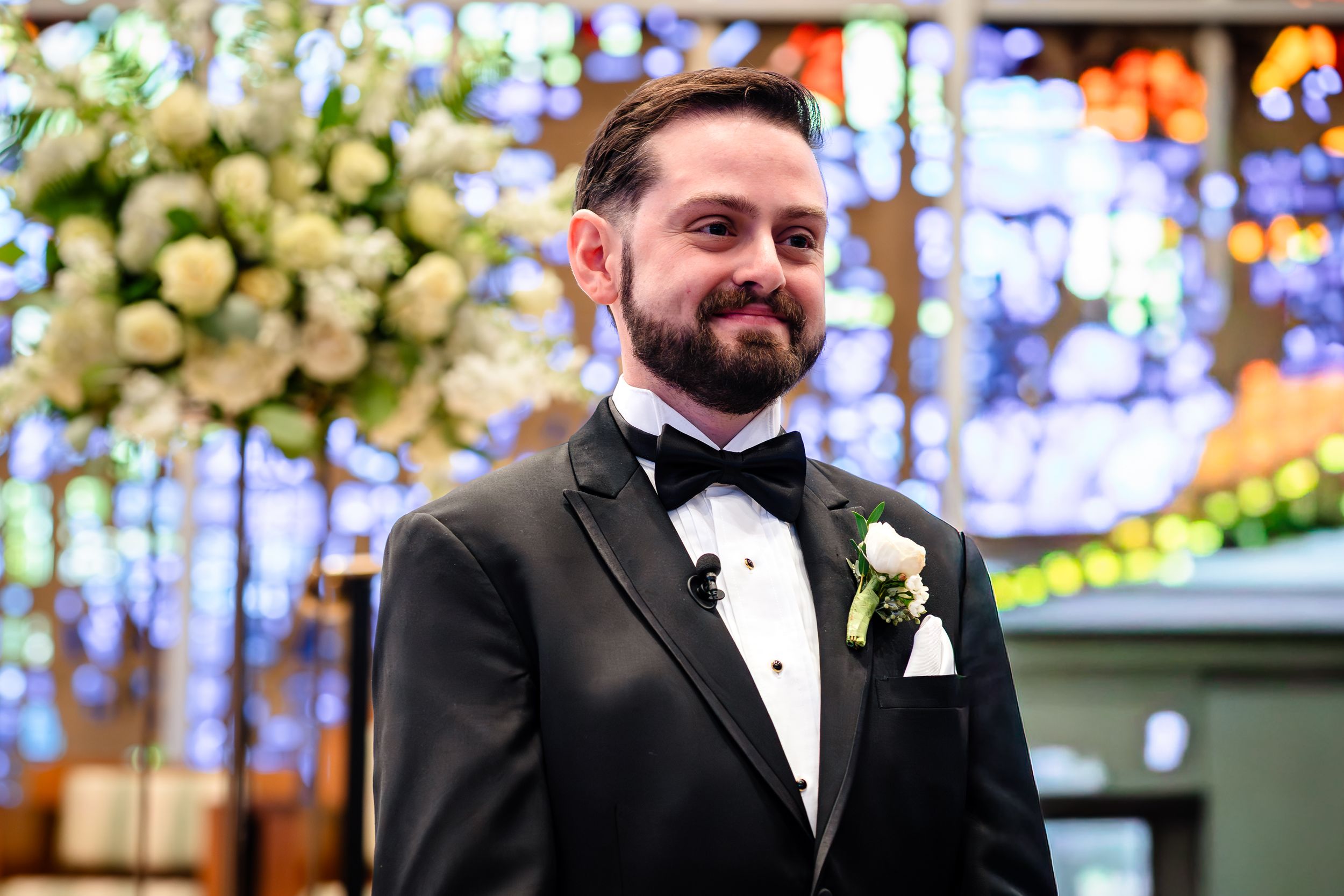 A groom in classic black tuxedo stands before colorful stained glass windows.
