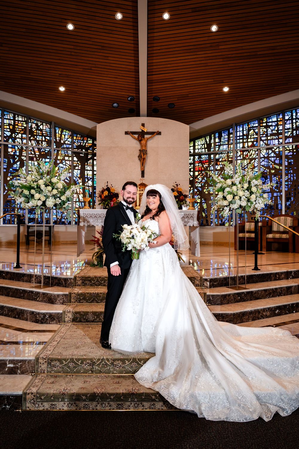 A wedding couple poses on church steps with elegant floral arrangements and stained glass windows.