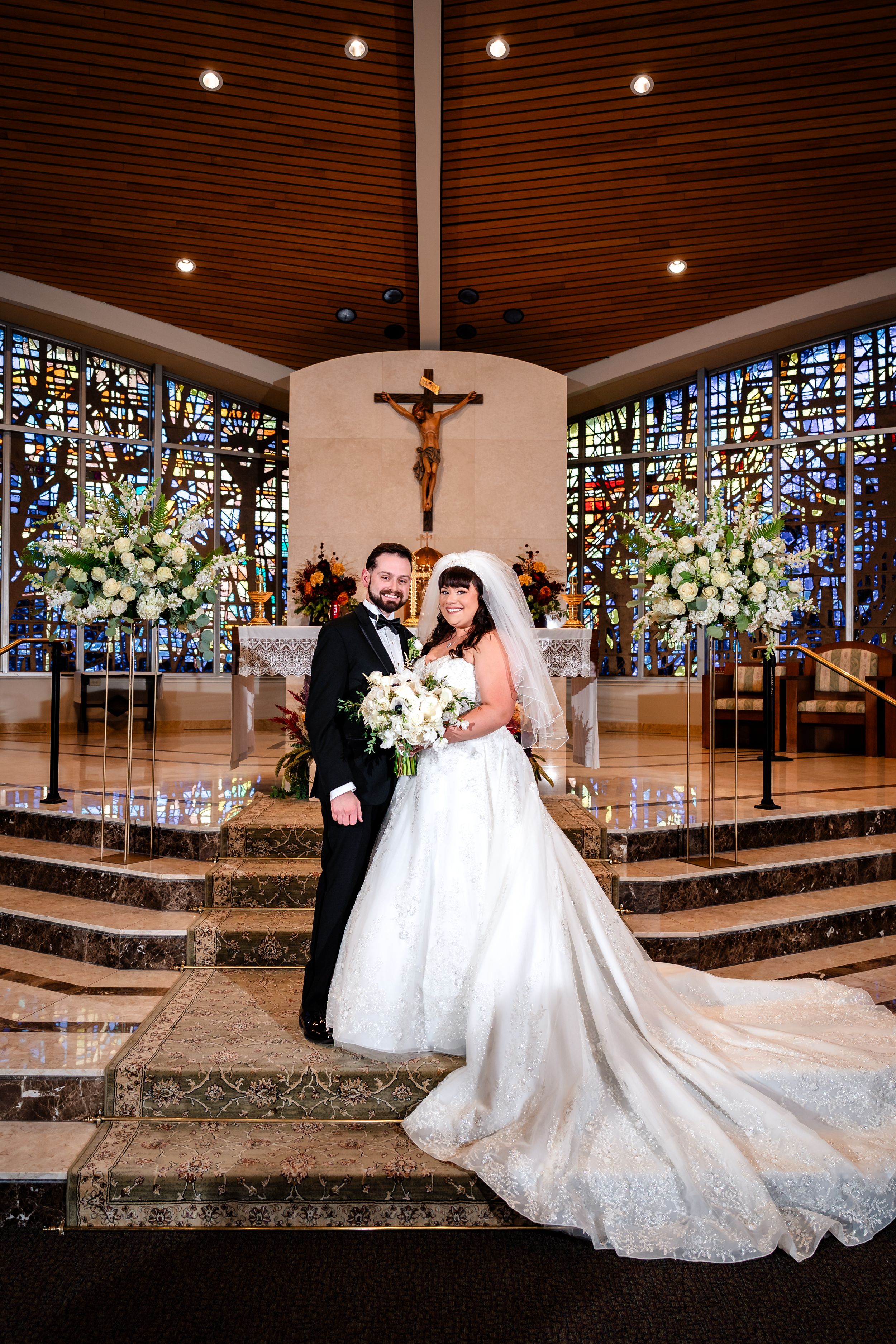 A wedding couple poses on church steps with elegant floral arrangements and stained glass windows.