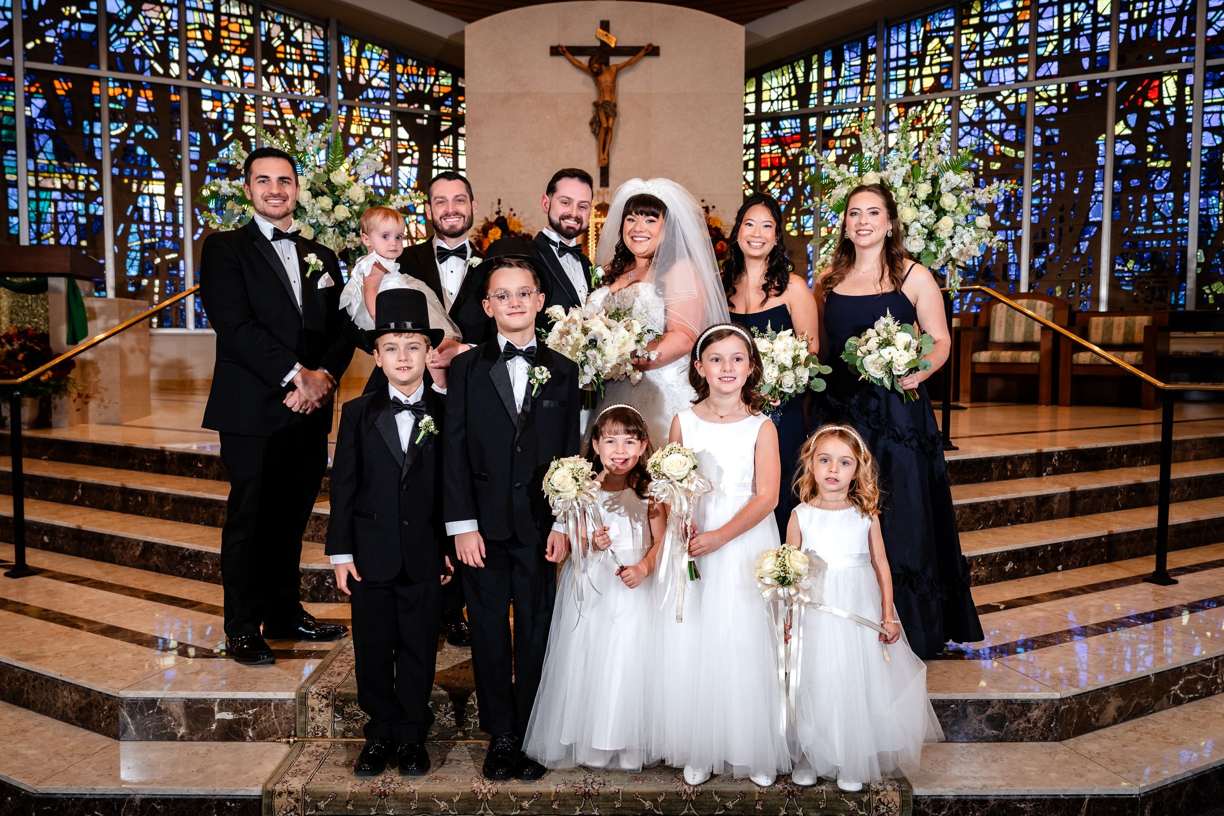 A formal wedding party poses together on church altar steps with floral arrangements.