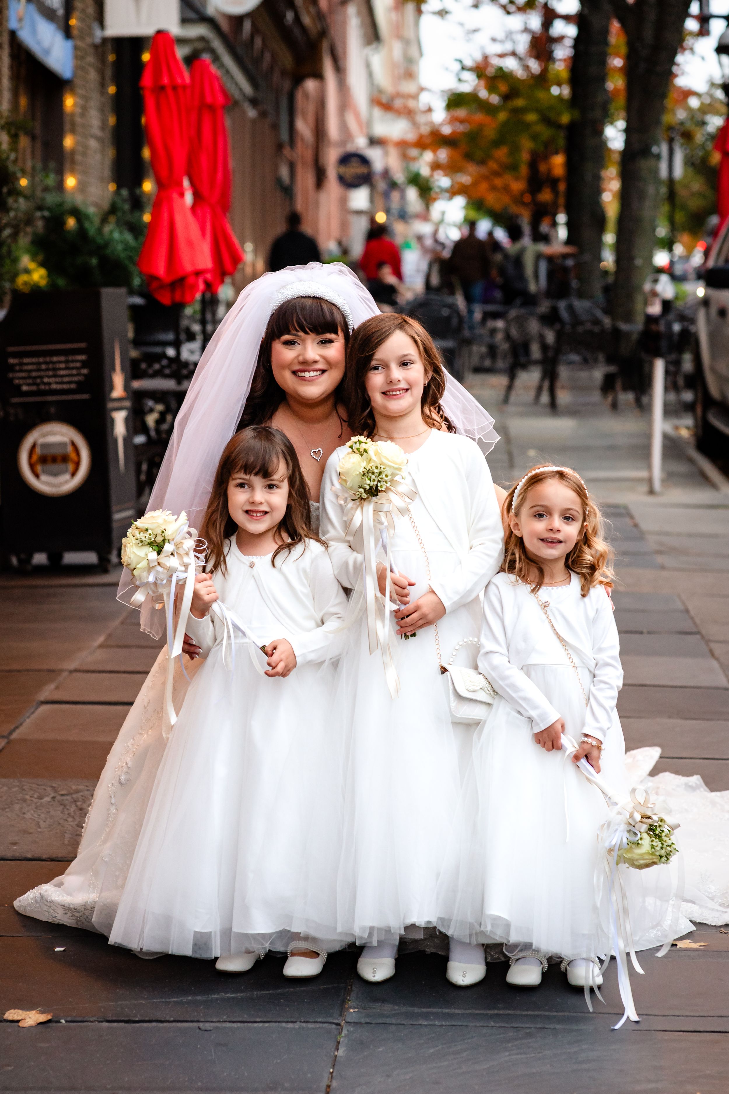 A bridal party in white dresses poses on a city sidewalk during autumn.